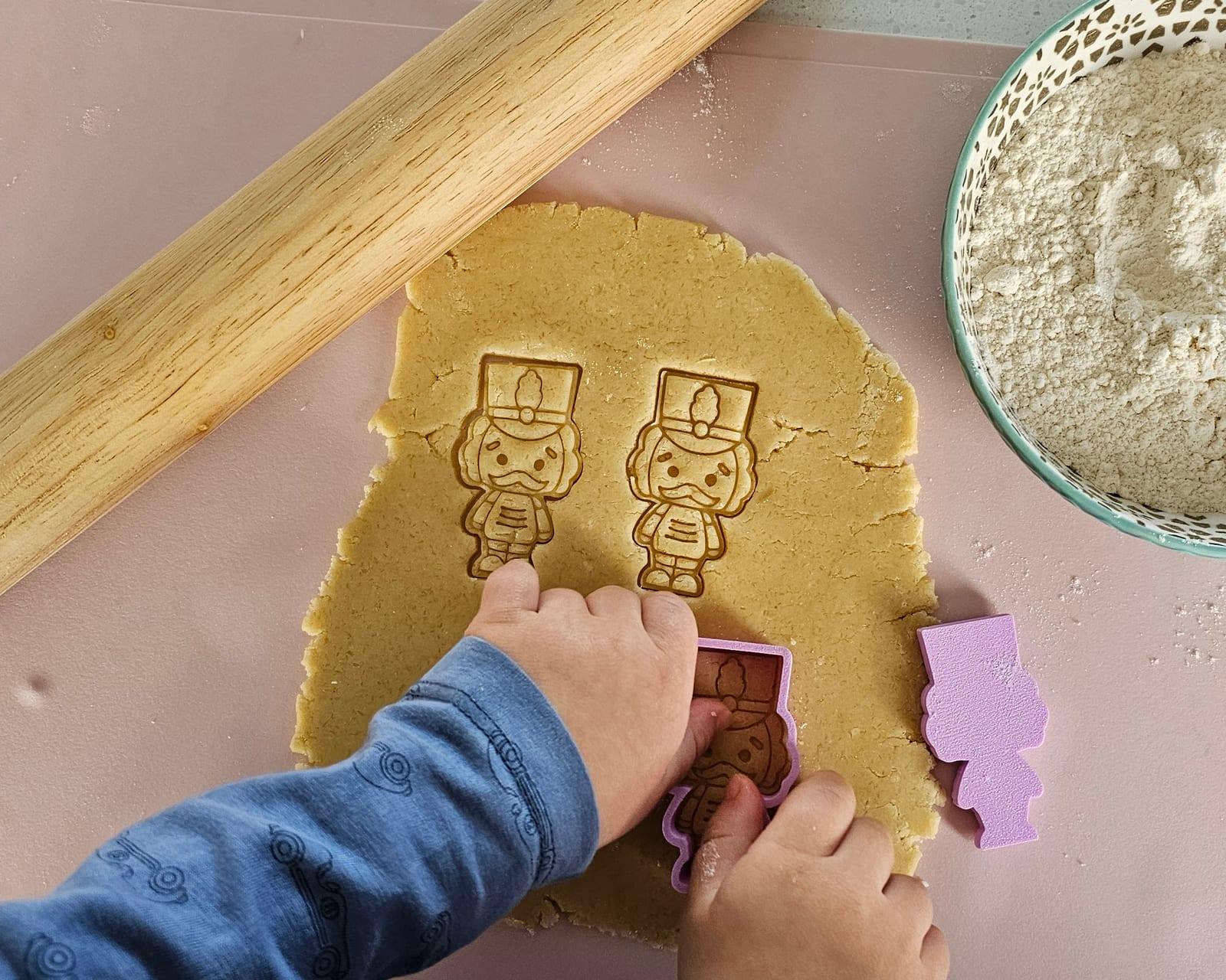 A child pressing a nutcracker cutter into rolled dough, beside a rolling pin and a bowl of flour